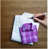 A hand is writing on a white shirt with a black marker, while a folded purple plaid shirt is placed on top of it. Produktbild-Vorschau