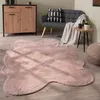 A pink, irregularly shaped rug is laid out on a wooden floor in a living room setting. A gray sofa with a throw blanket and a cushion is visible in the background, along with a wooden cabinet and a small side table with a plant. Voir les images du produit