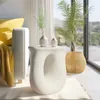A stylish white side table with a unique curved design, placed next to a sofa with a yellow cushion and throw, and a potted plant in a woven basket. The scene is set in a bright, modern living room with large windows offering a view of rolling hills. Náhled obrázku produktu