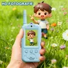 A hand holding a blue walkie-talkie with a cartoon boy displayed on its screen, with a larger cartoon boy figure in the background amidst grass and flowers. Produktbild-Vorschau