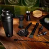 A collection of black barware tools laid out on a wooden table with a green tiled backsplash. Voir les images du produit