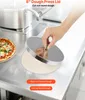 A hand is using a round metal lid to press dough on a stainless steel kitchen counter, with a pizza visible on the left. Podgląd zdjęcia produktu