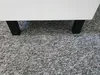 The bottom part of a white cabinet with black legs is shown resting on a grey carpet. Podgląd zdjęcia produktu