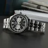 A close-up shot of a Seiko chronograph watch with a black dial and silver band, placed on a dark surface with blurred books and newspapers in the background. Náhled obrázku produktu