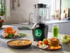 A blender is set up on a kitchen counter, surrounded by ingredients and prepared food. Náhľad obrázku produktu