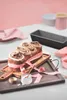A baked good, possibly a sweet bread or cake, is presented on a wooden board with pink ribbon and a small dish of what appears to be frosting or cream. The background is a soft pink and white, suggesting a styled setting. Náhľad obrázku produktu
