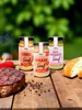 Three jars of mustard are displayed on a wooden table with a steak, bread, and chili peppers, suggesting a barbecue or meal setting. Produktbild-Vorschau