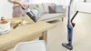 A person is using a handheld vacuum cleaner to clean crumbs off a wooden table. A standing vacuum cleaner is visible in the background. Voir les images du produit
