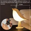 A bird-shaped lamp with a light emanating from its body, placed on a dark surface next to a couch. A close-up inset shows a hand adding essential oil to the lamp. Anteprima dell’immagine prodotto