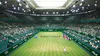 A tennis match is being played in a large, green stadium filled with spectators. The court is green and well-lit by overhead lights. Podgląd zdjęcia produktu