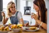 Two women eating breakfast at a table with a coffee maker in the background. Produktbild-Vorschau