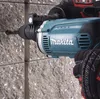 A close-up shot of a person wearing work gloves using a Makita hammer drill to drill into a concrete wall. Podgląd zdjęcia produktu