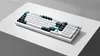 A white mechanical keyboard with black and teal keycaps is displayed on a white surface with a gray shadow. Produktbild-Vorschau