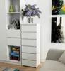 A white dresser with six drawers stands next to a white bookshelf filled with books and decorative items. A vase of purple flowers sits on top of the dresser. Produktbild-Vorschau