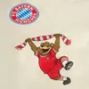 A cartoon bear mascot wearing a FC Bayern München jersey and scarf, with the team's logo above. Produktbild-Vorschau