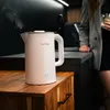 A white electric kettle is shown on a wooden cabinet in a modern kitchen setting, with a person holding a drink in the background. Produktbild-Vorschau