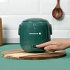 A hand is pressing a button on a green mini rice cooker placed on a wooden cutting board. Produktbild-Vorschau
