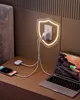 A bedside table with a laptop, phone, and charging accessories plugged into a wall outlet, illuminated by a glowing shield outline. Produktbild-Vorschau