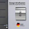 A modern, stainless steel mailbox is mounted on a light gray wall next to a dark gray door. The mailbox has three oval-shaped windows and a lock. A German flag map graphic with text is also present on the wall. Produktbild-Vorschau