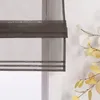 A close-up view of a dark gray sheer Roman blind with horizontal stripes, partially covering a window. A branch with artificial eucalyptus leaves is visible to the right. Produktbild-Vorschau