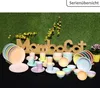 A collection of pastel-colored tableware, including plates, bowls, and cups, arranged on a black surface with a green, grassy background and decorative yellow flowers. Voir les images du produit