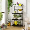 A multi-tiered black metal shelf unit filled with various potted plants and a yellow watering can, placed on a balcony or patio. Náhled obrázku produktu
