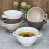 A collection of soup bowls with handles, some filled with soup, arranged on a textured surface with a wooden background and decorative plants. Produktbild-Vorschau