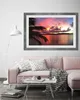 A living room scene with a pink sofa, a coffee table, and a framed picture of a sunset over the ocean hanging on the wall. Produktbild-Vorschau