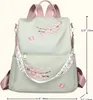 A light green backpack with pink cherry blossom embroidery and pink straps is shown against a white background. Dimensions are indicated with measurement lines. Náhľad obrázku produktu