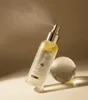 A bottle of d'Alba First Spray Serum is shown with mist spraying from the nozzle, next to a clear sphere on a gradient background. Náhled obrázku produktu