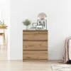 A wooden dresser with three drawers is placed against a white wall, with a lamp and framed pictures on top. A dining table and chair are visible in the background. Náhled obrázku produktu