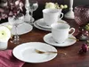 A beautifully set table with white porcelain dishes, gold cutlery, and crystal glasses, adorned with flowers. Produktbild-Vorschau