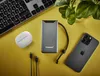A flat lay of tech accessories including a power bank, smartphone, wireless earbuds, and a keyboard, arranged on a yellow surface with a plant in the background. Produktbild-Vorschau