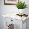 A close-up of a marble-patterned side table with a drawer and a shelf. A plant in a white pot, a black book, and a gold butterfly decoration are on top of the table. Books are on the shelf below. Produktbild-Vorschau
