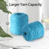 Two spools of blue yarn are shown on a white surface, with a glass of water and lemon in the background. Green leaves are in the foreground. Produktbild-Vorschau