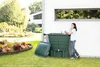 A woman is standing next to a green rain barrel in a garden, with a house in the background. Voir les images du produit