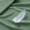 A close-up of a green textured fabric, likely a bedsheet, with a single white feather resting on it. Text overlays indicate softness and coziness. Anteprima dell’immagine prodotto