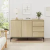A light wood sideboard with two doors and three drawers is shown in a well-lit room with a large window and a sofa. Voir les images du produit