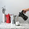 A hand is pouring coffee from a black Moka pot into a white cup, with two other Moka pots (red and silver) and a vase with dried flowers in the background. Anteprima dell’immagine prodotto