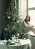 A woman sits by a window next to a decorated Christmas tree, with a table set for a festive meal in the foreground. Produktbild-Vorschau