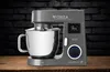 A kitchen mixer is displayed on a wooden table against a dark brick background. Produktbild-Vorschau
