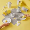 A breakfast setting with various tableware and food items arranged on a yellow and gray background. Anteprima dell’immagine prodotto