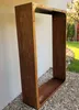 A rusted metal firewood rack is placed outdoors on a gravel surface next to a wall. Produktbild-Vorschau