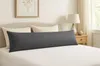 A long, dark gray body pillow is laid out on a white bed with a striped pattern. Two standard pillows are behind it, and a headboard is visible. A bedside table with a lamp and a decorative vase is to the right. Produktbild-Vorschau