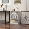 A white three-shelf bookcase is placed next to a desk with a lamp and a framed picture on the wall. Podgląd zdjęcia produktu