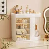 A white makeup organizer filled with various cosmetic products is displayed on a dresser, with a blurred plant and a framed picture in the background. Náhled obrázku produktu