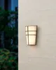 An outdoor wall lamp is mounted on a light-colored textured wall, with blurred greenery in the background. Produktbild-Vorschau