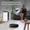 A woman is sitting on a couch, holding a remote control, while a robot vacuum cleaner is on the floor. A smartphone is also visible, showing an app interface. Náhled obrázku produktu