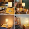Collage of four images showing a modern LED table lamp with a USB charging port in different settings: on a dining table, on a desk with a laptop, on a bedside table, and in a bedroom setting. Produktbild-Vorschau
