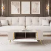 A modern living room with a beige tufted sofa and a beige high-gloss coffee table with gold legs. The coffee table has an open compartment revealing a gold geometric decor piece and a book. Produktbild-Vorschau